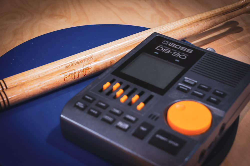 The Boss DB-90 metronome pictured with drumsticks, symbolizing its status as the best metronome for drummers who want a mix of technology and tradition for their practice sessions.