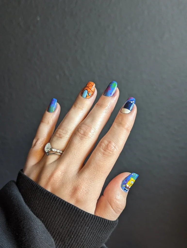Photo of a person's left hand with intricately designed nail art against a dark background. The thumb nail features a vibrant blue and purple design with white stars, resembling a galaxy.