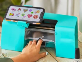 A person selects a nail design on their smartphone, which is attached to a bright turquoise nail art printer, demonstrating the ease of choosing digital nail art for personalized fashion statements.