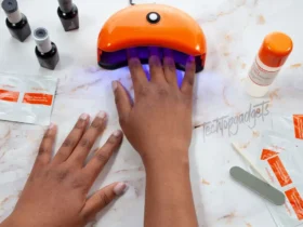 A hand placed under an orange nail dryer, with nail care products surrounding it, illustrating practical nail dryer tips and tricks for achieving top nail art trends.